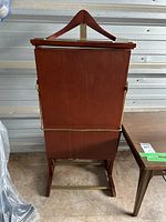 Front view of wood clothing valet showing hanger on top, brass support bars, and base frame, positioned against a storage unit wall.