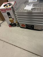 Hamilton Beach food dehydrator with trays stacked and Starfrit peeler box beside it on floor.