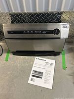 Front and top view of the stainless steel FoodSaver V3840 vacuum sealer with Quick Start Guide and User Manual on floor in front of it.