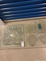 Set of various clear glass serving platters and plates arranged on floor including large square divided platter, two triangular plates, a rectangular plate, and a small green glass pedestal.