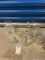 Six assorted clear glass items on a surface: two pitchers, one tall pitcher with swirl pattern, two lidded jars, one sauce boat