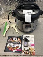 Front and top view of Instant Pot Duo stainless steel electric pressure cooker with digital display, showing control buttons and brand logo. Positioned next to instruction manual, recipe booklet, and three spoons in different colors.