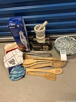 Overview of kitchen lot items laid on floor: Oster hand blender in box, Black+Decker mini chopper in box, white marble mortar and pestle, wooden utensils, kitchen towels, enamel colander, whisk, and crochet pot holder.