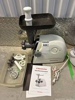 Photo showing Sunmile electric meat grinder with metal grinding attachment mounted, white plastic plunger, two grinding plates, sausage stuffing funnels, clear plastic tray with accessories, electrical cord, and instruction manual laid out on surface.