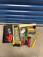 Painting and hardware tools arranged showing paint rollers, paint tray, hacksaw, sanding block, flashlights, and screws