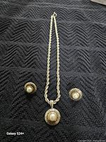 Photo displays the full necklace with rope chain and matching earrings laid flat on a dark textured surface, showing design details.