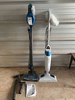 Full view of Shark HV300 Rocket stick vacuum and Bissell steam cleaner side by side with attachments and manual on floor