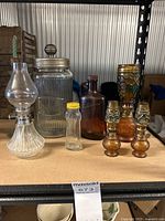 View of assorted vintage glassware including oil lamps, large jar with metal lid, small jar with yellow lid, decorative candle holders, brown bottle, and floral decorated tall jar.