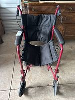 Front view of foldable transfer wheelchair showing red frame, black fabric seat and backrest, and black padded armrests