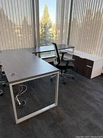 Office desk set consisting of a rectangular desk, L-shaped return desk, and two-drawer filing cabinet under large windows with vertical blinds.