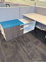 Wide shot of cubicle corner with white laminate desk, grey fabric cubicle panel, and pedestal storage drawers topped with blue cushion.