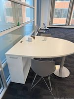 Side view of white side desk table showing semi-circular tabletop, single pedal support leg, and attached storage cabinet with handle. Gray chair with metal legs is positioned in front.