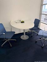 Overall view of the white semi-circular side desk table with 3 blue office chairs in a room with white walls and window.