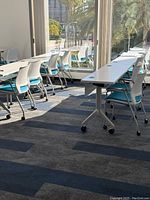 Training tables arranged in rows with white mesh chairs and wheels visible on table legs, sunlight through windows.