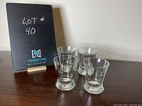 Four clear glass cups with raised bee design on the side and thick base, arranged on dark surface with lot label in background.