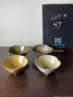 Four handmade ceramic bowls in various colors and glazes displayed on dark wooden table with LOT #47 sign in background