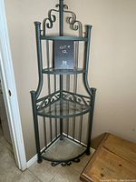 Front view of glass and metal corner shelf with ornate scrollwork design, three glass shelves, tall and narrow shape fitting into corner space.