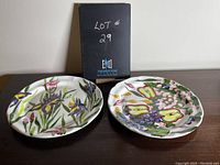 Two hand-painted ceramic serving plates by Hugo Quattrocchi displayed side by side showing colorful botanical and fruit motifs.