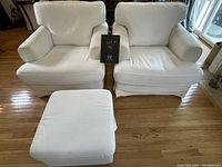 Two large white IKEA chairs with removable slipcovers and matching white storage ottoman on hardwood floor near glass door.
