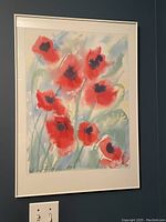 Framed watercolor print showing vibrant red poppies, signed by Linda Fay Powell, hanging on a wall with light switch visible below.