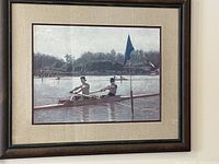 Framed print showing two men rowing in a shell on a water body with trees in the background, framed with cream colored matting and brown wooden frame. Minor nicks are visible on the frame.