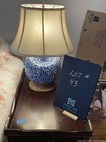 The lamp placed on a dark wooden side table next to a sofa, turned on showing the beige lampshade and blue and white ceramic base with pattern.