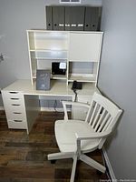 Front and side view of white office desk with attached shelving, filing cabinet drawers on right, and white office chair beside it.