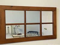 Front view of a rectangular wood framed mirror divided into six panes like a window reflecting part of a room including a chair and door.