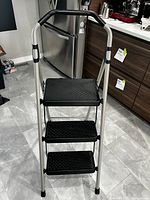 Front view of 3 step aluminum ladder showing three black textured non-slip steps and curved top handle with black grips.