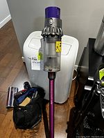 Assembled cordless vacuum standing upright showing purple cyclone head, clear bin and attached purple wand with black cloth bag containing attachments on floor