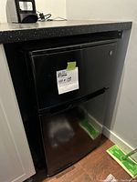 Frontal view of black GE mini fridge under countertop showing freezer top door and fridge bottom door with lot label.