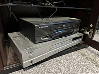 Photo shows two electronic players stacked inside a cabinet: KOSS silver DVD player on bottom and Electrohome black VHS player on top.