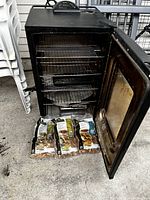 Front view with door open showing three interior racks, drip pan, water pan and three wood chip bags in front.