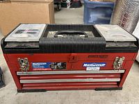 Front view of the Mastercraft red metal tool box showing two drawers and latches, with parts organizers on the black lid.