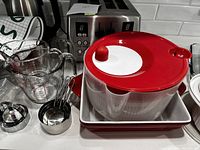Photo showing clear glass measuring cups, stainless steel measuring spoons, red salad spinner with lid and colander, and red rectangular stoneware baking dish.