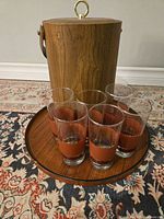 Photo showing six cylindrical glasses with burnt orange bands and a wooden round tray holding them, and wood grain ice bucket behind