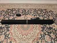 Full front view of black Cowin soundbar with power adapter and remote placed on patterned rug.