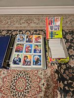 Blue 3-ring binder opened to hockey trading cards with nine per page, accompanied by a yellow and colorful Pro Set 1990/91 EPL soccer card box full of cards.