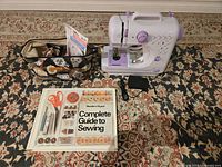 Overview of the sewing machine, storage bag with accessories, power adapter, foot pedal, and sewing guide book laid out on a patterned carpet.