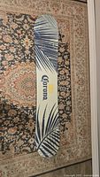 Top side of snowboard showing cream background with dark blue palm frond graphics and Corona logo