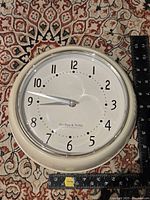 Front view of the cream-colored Sterling & Noble wall clock with black numbers and silver hands placed against patterned fabric, rulers show approximately 8 inches diameter.
