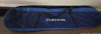 Full view of the Dakine snowboard bag showing two-tone blue fabric, front pocket with logo, and black carrying straps.