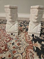 Pair of white marble moai bookends photographed on patterned rug, showing front and side views highlighting angular carved features and robe details