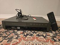 Front view of Mitsubishi HS-U341 VHS player with cables on top and remote control beside it.