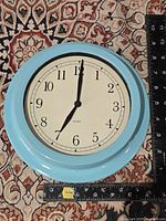 Front view showing robin's egg blue frame, cream dial with black numbers and black hands, ruler beside clock indicating about 9-inch diameter.