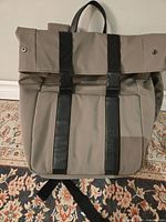 Front view of gray canvas backpack with black leather vertical strips and roll-top secured with black buckles.