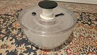 Salad spinner fully assembled, showing clear plastic bowl, clear lid with black push button and locking lever