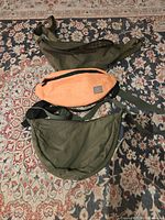 Three adjustable waist fanny packs placed on a patterned carpet: two olive green and one orange in the middle. All have zippered closures and adjustable straps.