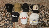Overview photo showing all seven hats displayed on a rug: two Titleist golf hats (black and white/red) in front row and other five hats in back row