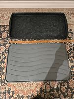 Two rectangular rubber mats side by side on patterned carpet: one black with raised circular bumps, one gray with wave pattern. Both have raised lip edges.